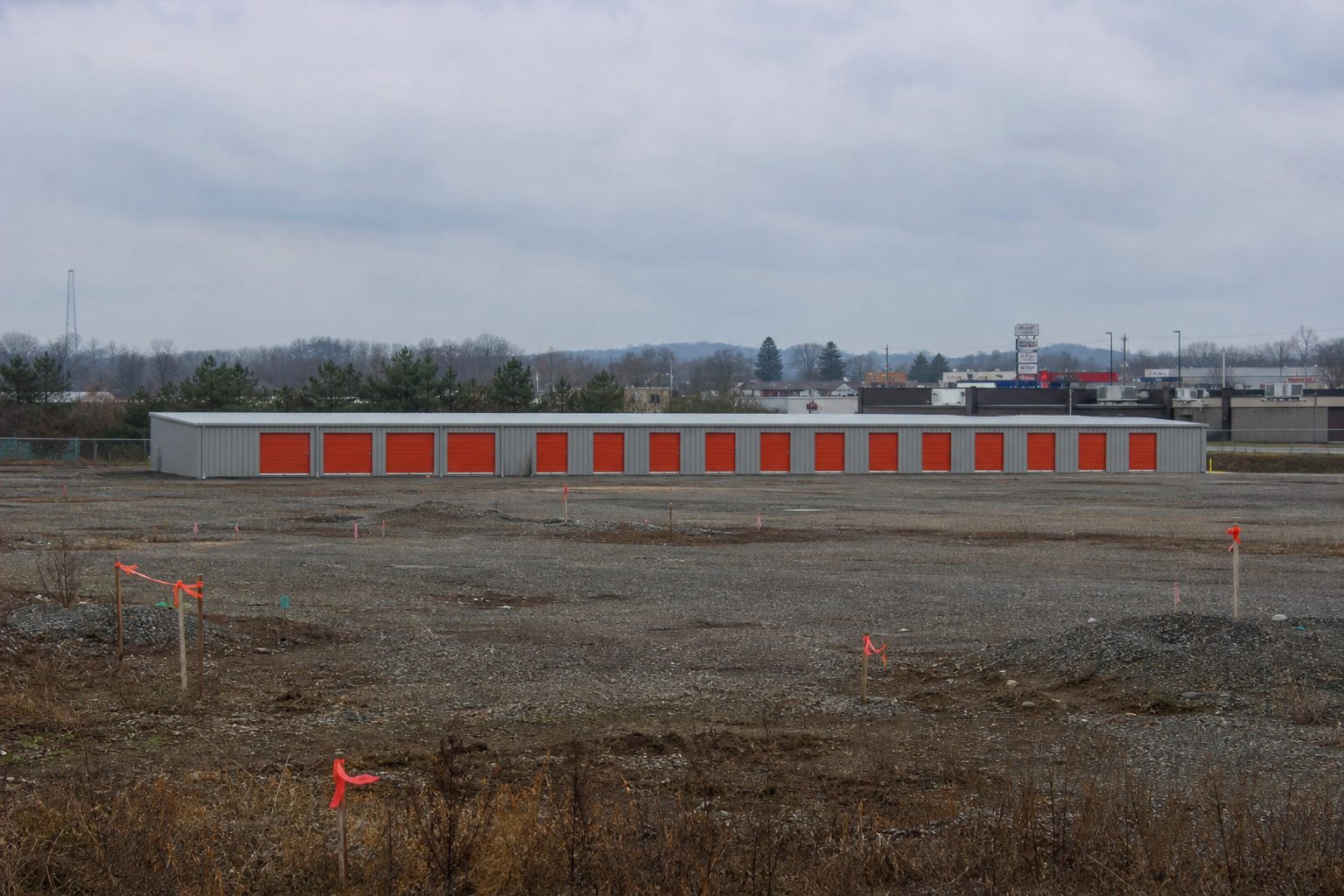 Single story storage building across an empty gravel lot with survey stakes standing in the foreground